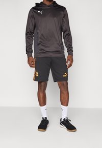 Czarna bluza z kapturem Puma z białym logo, zestawiona z szarymi szortami z pomarańczowymi akcentami i logo. Białe skarpetki i czarne buty dopełniają cały look.