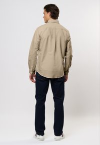 Uomo in piedi di spalle, vestito con una camicia beige a maniche lunghe e bottoni, jeans blu scuro e sneakers bianche con strisce rosse e blu.