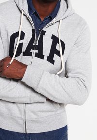 Felpa grigia con zip e grande logo nero "GAP" sul petto, dotata di cappuccio e lacci, realizzata in morbido cotone.