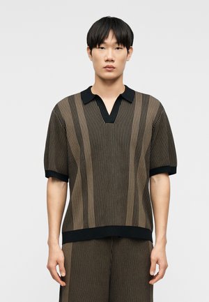 Junger Mann mit glattem schwarzem Haar, der ein kurzärmliges Hemd mit schwarzen und braunen vertikalen Streifen sowie passende Hosen trägt und vor hellem Hintergrund steht.