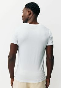 Muž s krátkymi kučeravými vlasmi otočený chrbtom, oblečený v jednofarebnom bielom krátkornom tričku a béžových nohaviciach na bielom pozadí.