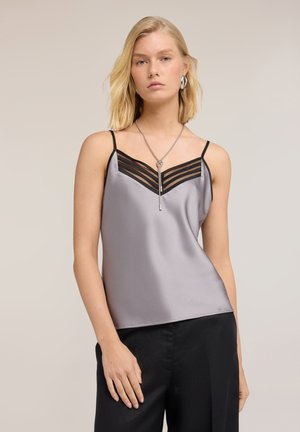 Blonde vrouw die een zilveren satijnen camisole draagt met zwarte mesh-detail en zwarte broek, geaccentueerd met zilveren oorbellen en een lange ketting.