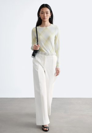 Vrouw met lang donker haar, een licht geruit shirt met lange mouwen, witte wijde broek met hoge taille, zwarte sandalen en een zwarte schoudertas.