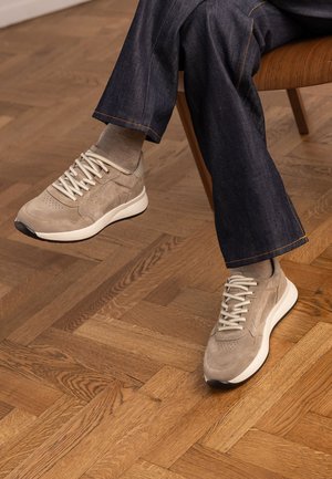 Person wearing beige suede sneakers and light brown socks, seated on a wooden chair, with dark blue jeans and parquet flooring.