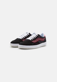 Baskets en daim noir avec une semelle en caoutchouc blanc, présentant des accents de couture rouges et des lacets blancs. Le logo Vans est affiché sur la languette.