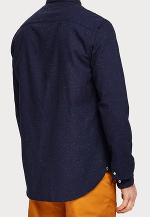 Man draagt een donkerblauw gemêleerd shirt met lange mouwen en mosterdgele broek, gezien van achteren met de rechterarm licht gebogen.