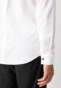 Férfi fehér, hosszú ujjú ingben fekete mandzsettával és fekete nadrágban, keze az oldalán pihen.