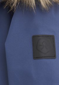 Blått tyg med en strukturerad yta. Har en svart rektangulär platta med en emblem, omgiven av beige fuskpäls upptill.