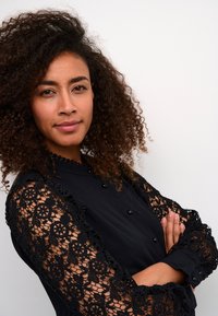 Black blouse featuring intricate lace sleeves with floral patterns, button details, and a smooth fabric body. Model displays arms crossed.