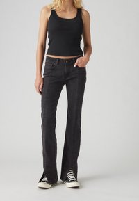 Pantalon flare en denim noir avec fentes latérales et coutures contrastées. Associé à un débardeur noir et des baskets noires.