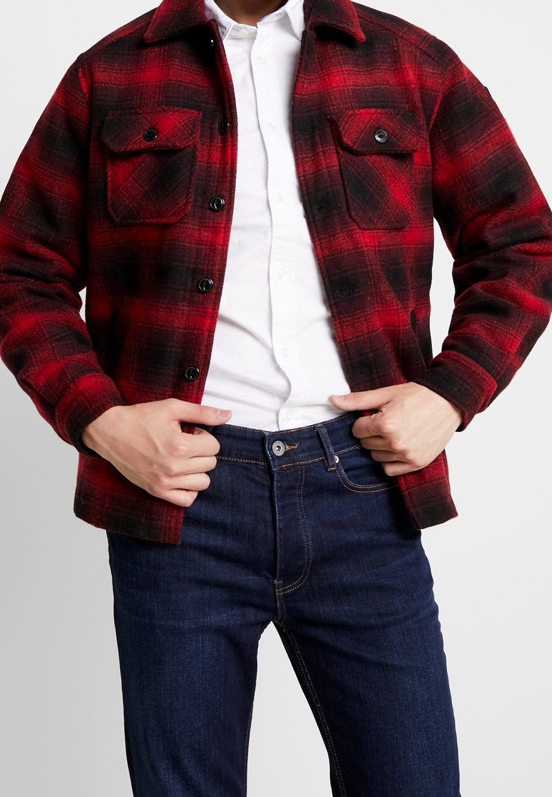 Veste en flanelle à carreaux rouges et noirs avec deux poches poitrine, boutons noirs et col de chemise, portée sur une chemise blanche à boutons.