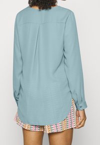 Halenka s dlouhým rukávem z měkké, světle modré látky s kulatým výstřihem a zadním skladem; doplněná barevnými vzorovanými šortkami.