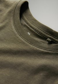Olivgrünes T-Shirt mit geripptem Rundhalsausschnitt und glatter Baumwolltextur. Kleines Etikett innen mit weißem Text.