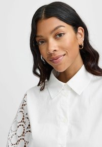 White button-up shirt with a classic collar, featuring intricate lace details on the sleeves. Smooth fabric with a relaxed fit.