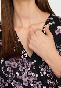 Robe noire à motifs floraux violets et blancs, design à col en V, avec une main délicatement posée sur le décolleté. Texture de tissu lisse.