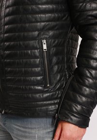 Giacca in pelle nera con design trapuntato e tasche laterali con zip. Caratterizzata da una texture liscia e una vestibilità slim; accenti in metallo della cerniera ben visibili.
