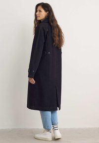 Svart denim lång kappa med slagkrage, sidoknappar och slits baktill, kombinerad med ljusblå jeans och vita hög-top sneakers.