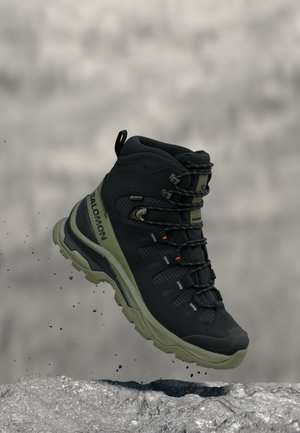 Scarpa da trekking Salomon nera e verde sospesa in aria sopra un terreno roccioso, vista di profilo laterale con suola robusta e ganci per i lacci.