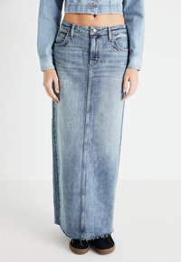 Gonna lunga in denim di colore azzurro con taglio dritto, cuciture frontali e orlo sfrangiato; abbinata a scarpe scure.