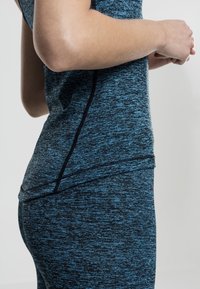 Gros plan d'une personne portant un haut de sport et un legging à motifs bleus et noirs, les mains jointes devant, debout devant un fond uni.