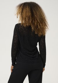 Top lungo maniche in pizzo nero con motivo floreale, dotato di scollatura rotonda e materiale testurizzato. Indossato con pantaloni neri.