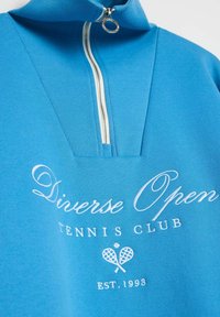 Niebieski sweter z wysokim kołnierzem i pół zamkiem. Posiada białe haftowane napisy "Diverse Open Tennis Club" oraz wzór krzyżujących się rakiet.