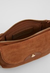 Bruine suède tas met een gouden ritssluiting; het interieur heeft een zachte voering en een label. De sluiting wordt beveiligd met een ronde metalen drukknoop.