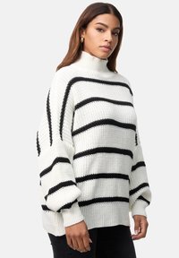 Pull en grosse maille avec rayures horizontales noires sur fond crème, col montant et poignets côtelés. Tissu texturé et coupe décontractée.