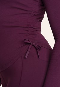 Lange mouwen top in diep paars, met gathers aan de zijkanten en een strikaccent op de taille. Gemaakt van een zachte, rekbare stof.