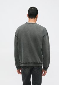 Sweatshirt cinza escuro com punhos e barra canelados, apresentando costuras com bordas crus e um corte descontraído. O tecido parece suave com um aspecto ligeiramente usado.