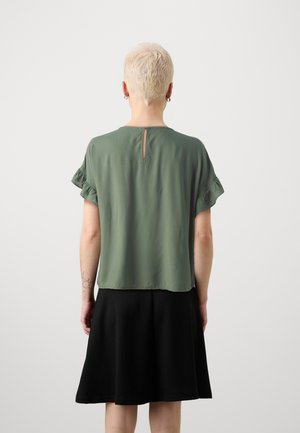 Personne aux cheveux blonds courts portant un chemisier vert à manches courtes avec des manches volantées et une jupe noire longueur genou, vue de dos.