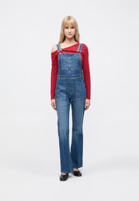 Salopette di jeans blu con gambe larghe e spalline regolabili, abbinata a una maglia rossa a maniche lunghe. Tessuto liscio, tasche frontali visibili.