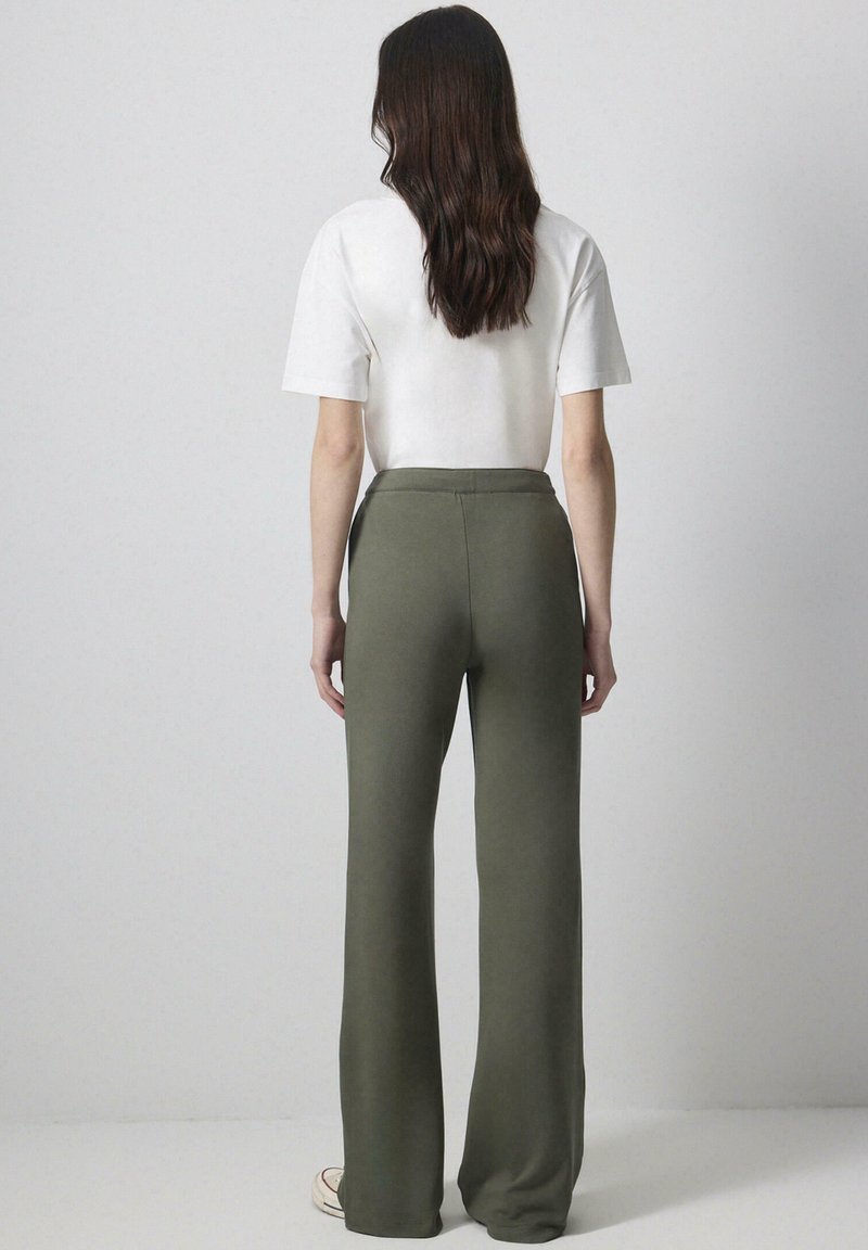 Γυναίκα με μακριά σκουρόχρωμα μαλλιά, που στέκεται με την πλάτη προς τα εμπρός, φορώντας λευκό κοντομάνικο πουκάμισο, χακί φαρδιά παντελόνια και λευκά παπούτσια.