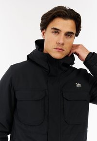 Juodas, vandeniui atsparus švarkas su aukštu apykakliniu, dviem krūtinės kišenėmis ir elastingais rankogaliais; turi mažą logotipą kairėje krūtinės pusėje.