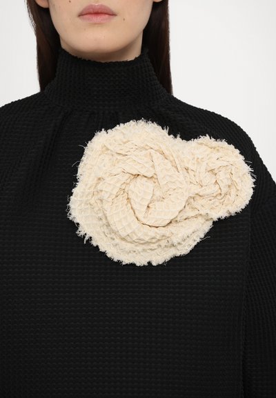 Maglione nero dalla texture con una grande spilla in tessuto color crema annodata, caratterizzata da un motivo arricciato e bordi sfrangiati, posizionata sul lato sinistro.
