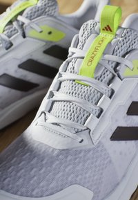 Szare buty sportowe z siateczkową cholewką, czarnymi akcentami, jaskrawą żółtą metką na języku i teksturowaną podeszwą. Wyposażone w tradycyjny system sznurowania.