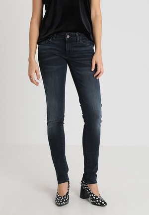 Jeans ajustados de mezclilla en un tono azul oscuro, con cierre de botón en la parte delantera y diseño de cinco bolsillos, combinados con zapatos de tacón alto negros con lunares blancos.