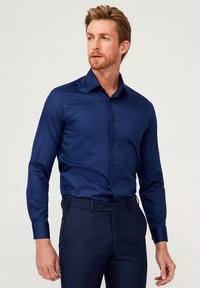 Donkerblauwe langekorte shirt met een strakke, gladde stoftextuur en een knoopsluiting kraag, gecombineerd met donkere, op maat gemaakte broeken.