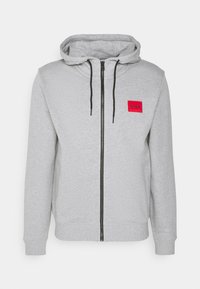 Sudadera con cremallera gris hecha de una tela texturizada, con cremallera negra, cordones ajustables y un parche rojo con el logo de la marca.