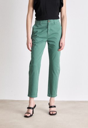 Personne portant un pantalon vert taille haute à jambes droites et des sandales noires à talons, debout devant un fond clair uni.