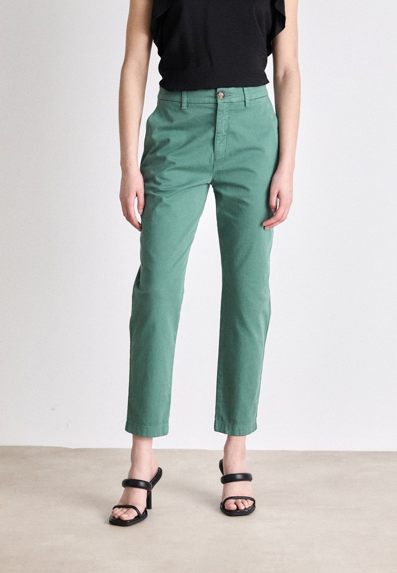 Personne portant un pantalon vert taille haute à jambes droites et des sandales noires à talons, debout devant un fond clair uni.