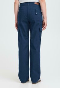 Person med marineblå cargo-bukser med klaplommer, knapdetaljer og beige sneakers, som står foran en ensfarvet lys baggrund.