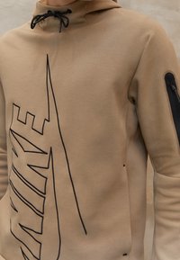 Beige hoodie med svarta dragsko och stor svart konturerad Nike-logga över bröstet; dragkedjeficka på vänster ärm.