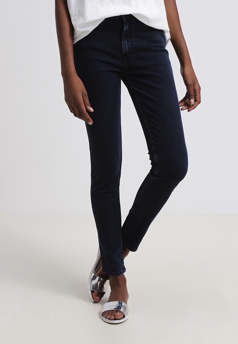 Una persona lleva puestos unos pantalones ajustados oscuros y sandalias plateadas, de pie sobre un fondo neutro, mostrando un atuendo casual.