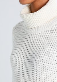 Weißer gestrickter Rollkragenpullover mit einem strukturierten, offenen Gewebemuster. Der Kragen ist gerippt und bietet einen deutlichen Kontrast in der Textur.