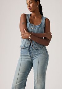 Γυναίκα με πλεξούδες, φορώντας αμάνικο τζιν γιλέκο με κουμπιά και τζιν παντελόνι, με σταυρωμένα τα χέρια, δείχνοντας τατουάζ εντόμου στον άνω βραχίονα και ασημένια κοσμήματα.