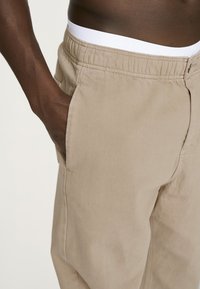 Beige bomuldsbukser med elastisk talje, sidelommer og en afslappet pasform, med synlige syninger.