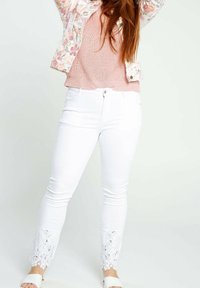 Jeans bianchi skinny con dettagli in pizzo sui polsini, abbinati a un top rosa in maglia e una giacca con motivo floreale.