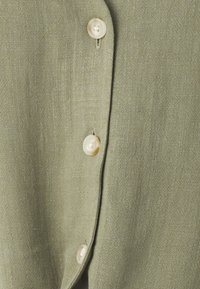 Tessuto di lino verde oliva con una leggera trama e una lucentezza discreta, caratterizzato da bottoni rotondi di colore chiaro lungo il pattina anteriore.