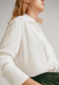 White long-sleeve blouse with a pleated accent along the button placket, featuring a standard collar and soft texture.